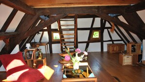 A cozy attic room with wooden beams, a red chair, a wooden table with a vase of flowers, and a ladder leading to the upper floor.