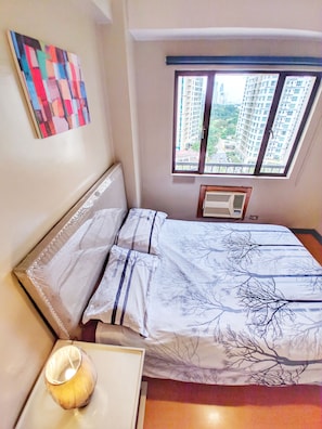 A bedroom with a bed, a bedside table with a lamp, a window with a view of buildings, and a wall-mounted air conditioner. - Forbeswood Heights (Taguig)
