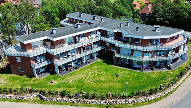 En moderne bygning med flere etager, buet tag, store glasfacader og udendørs siddepladser.