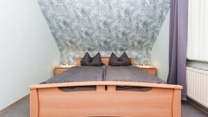 Una cama con estructura de madera y dos almohadas, una mesita de noche con una lámpara, y un papel tapiz estampado.