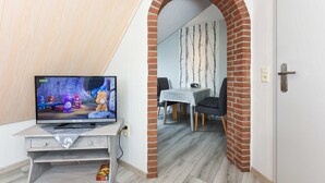 Una habitación con una televisión que muestra personajes animados, una zona de comedor con una mesa y sillas, y un arco de ladrillo que conduce a otra habitación.