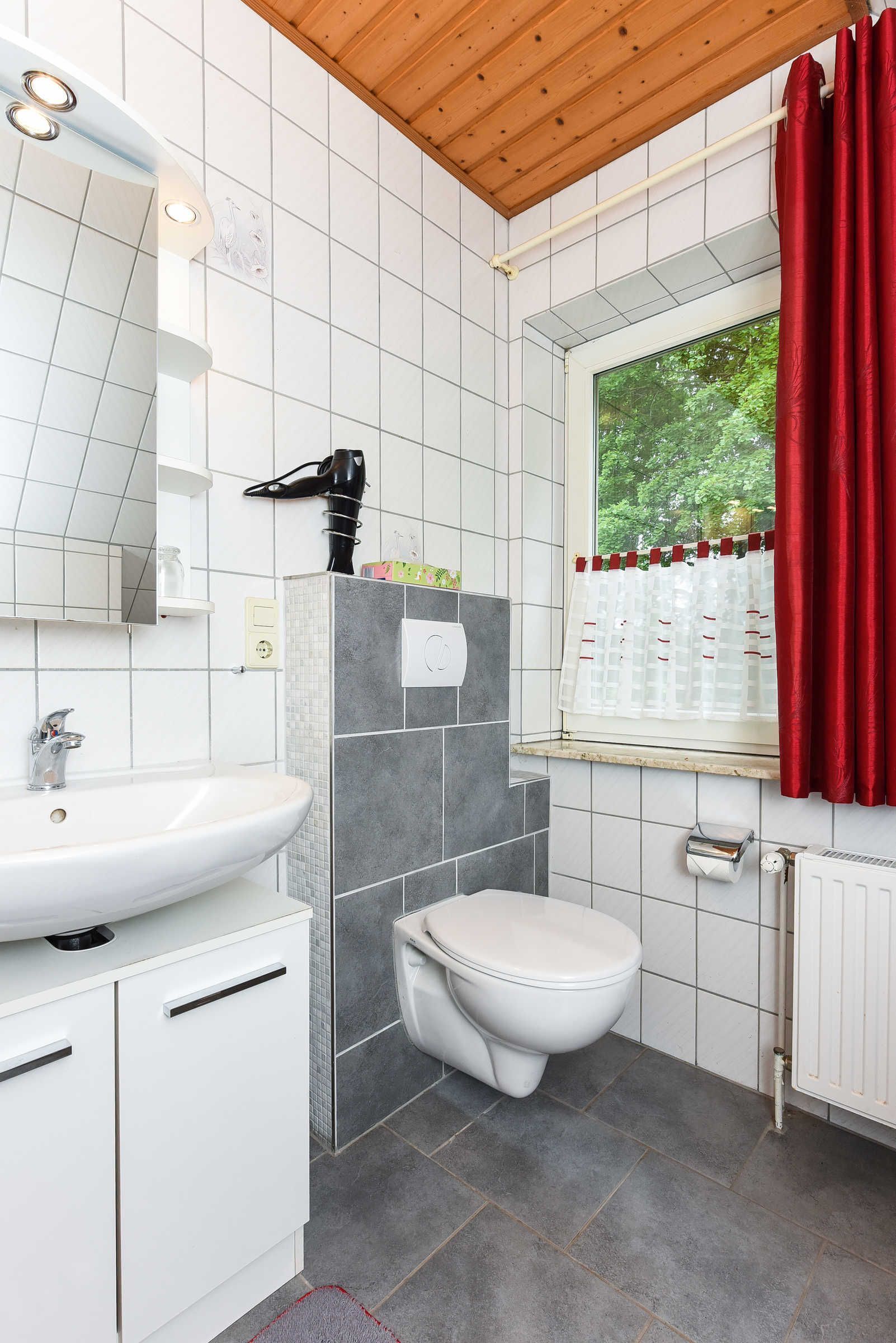 Une salle de bain avec un lavabo blanc, des toilettes et une fenêtre avec des rideaux rouges.