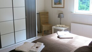 Ein Schlafzimmer mit Bett, Sessel, kleinem Tisch mit Lampe, einem Bild an der Wand und einem Fenster mit Blick auf Grünflächen.