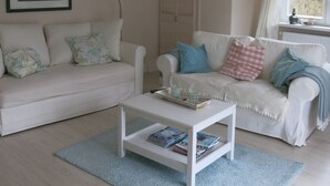 Ein Wohnzimmer mit zwei weißen Sofas, einem Couchtisch, einem gerahmten Bild an der Wand und einem Fenster mit Vorhängen.