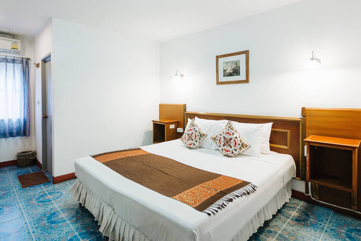 Hotellihuone, jossa on suuri sänky, kaksi yöpöytää, yölamppu, ikkuna verhoilla, seinälle ripustettu taulu ja kattovalaisin.