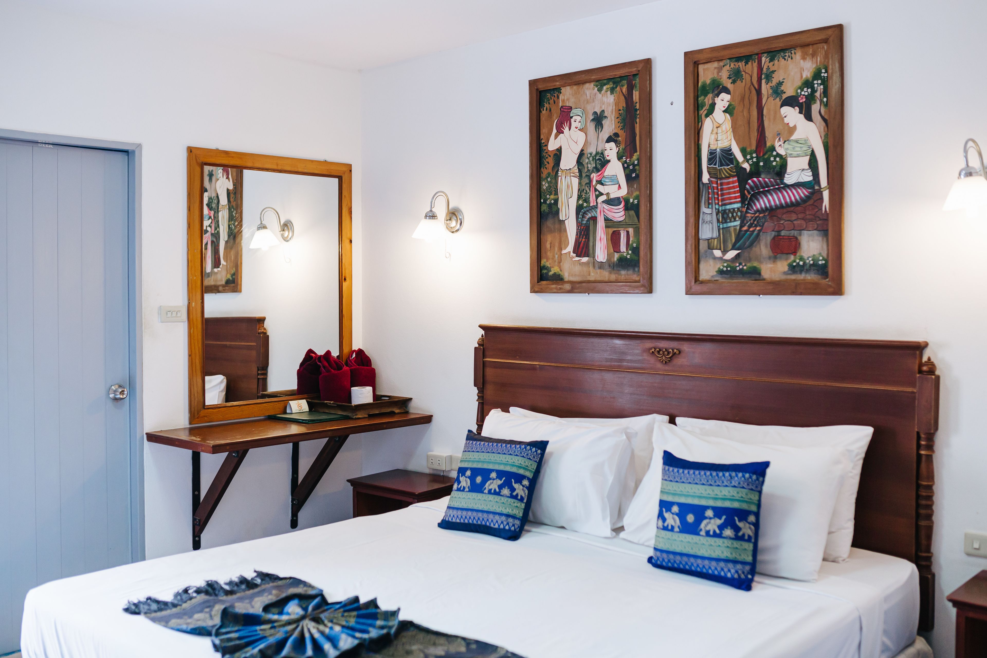A neatly made bed with white linens and patterned pillows, a wooden nightstand with a red bag, a wooden headboard, and two wall-mounted paintings.