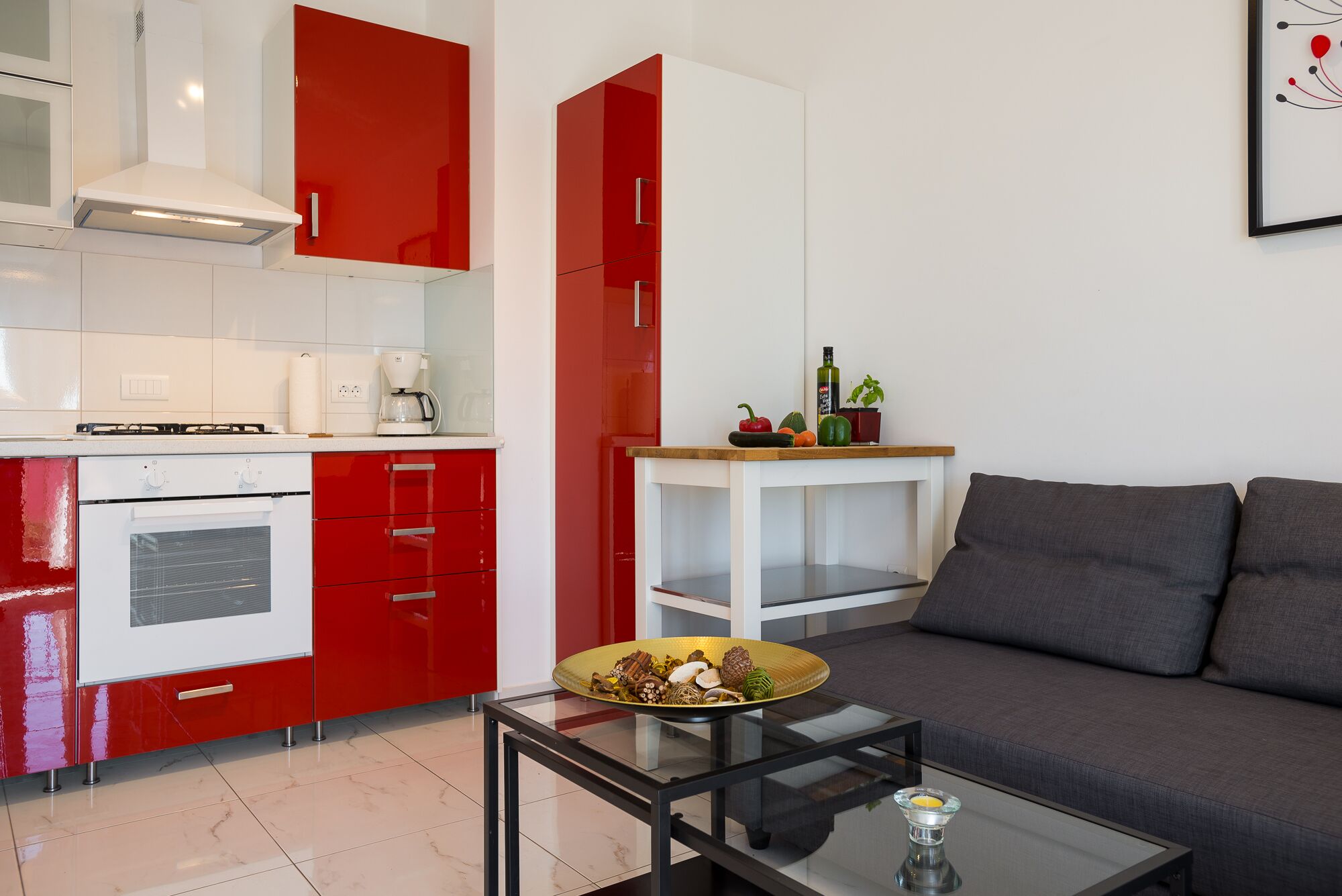 A modern kitchen with red and white cabinets, a built-in oven, and a coffee maker. There is a glass table with a bowl of fruits and a candle. A grey sofa and a white side table with a plant are visible in the living area.