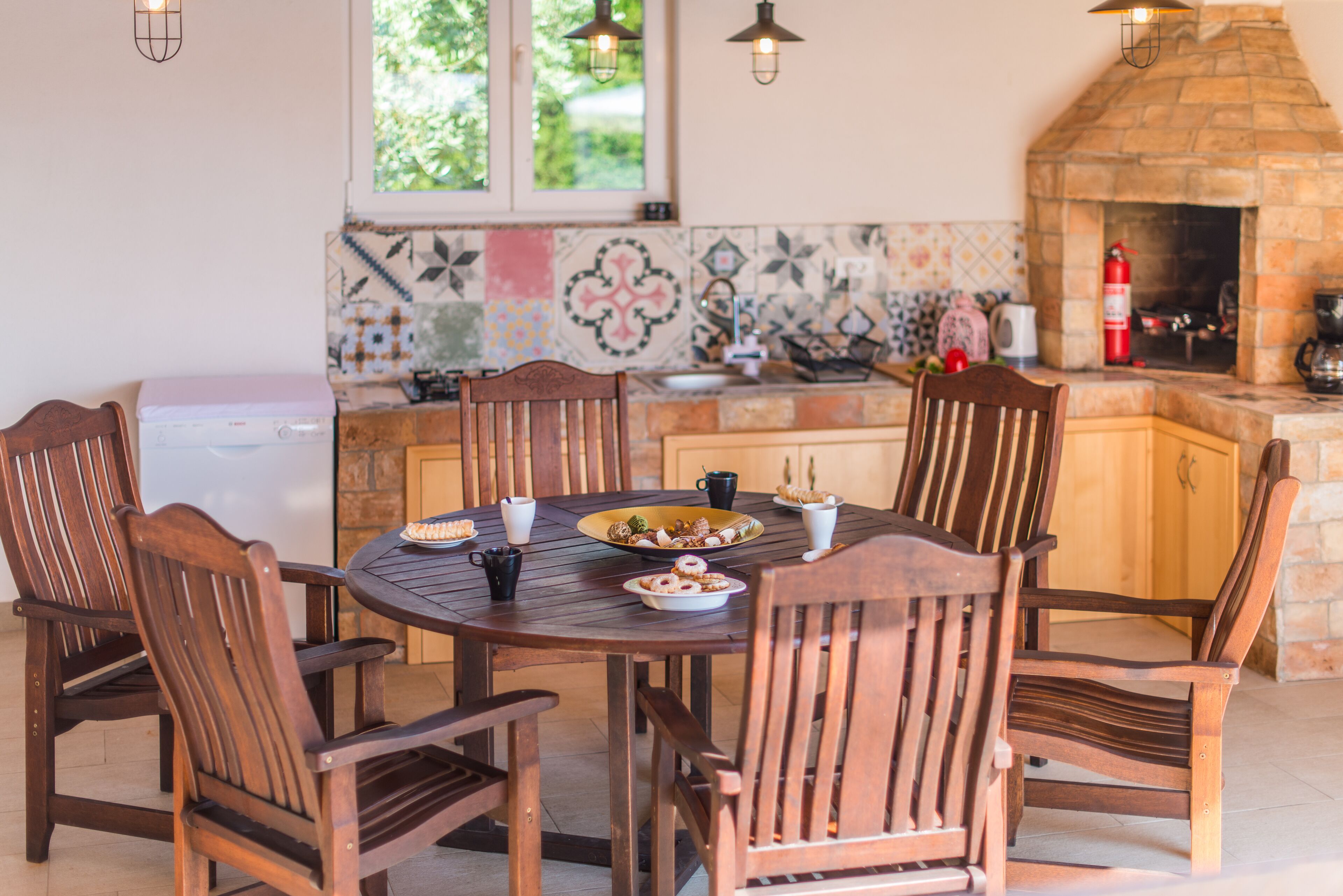 Un coin repas avec une table ronde en bois et six chaises en bois. Le mur derrière le comptoir est recouvert de carreaux, et on y trouve également une cheminée en pierre.
