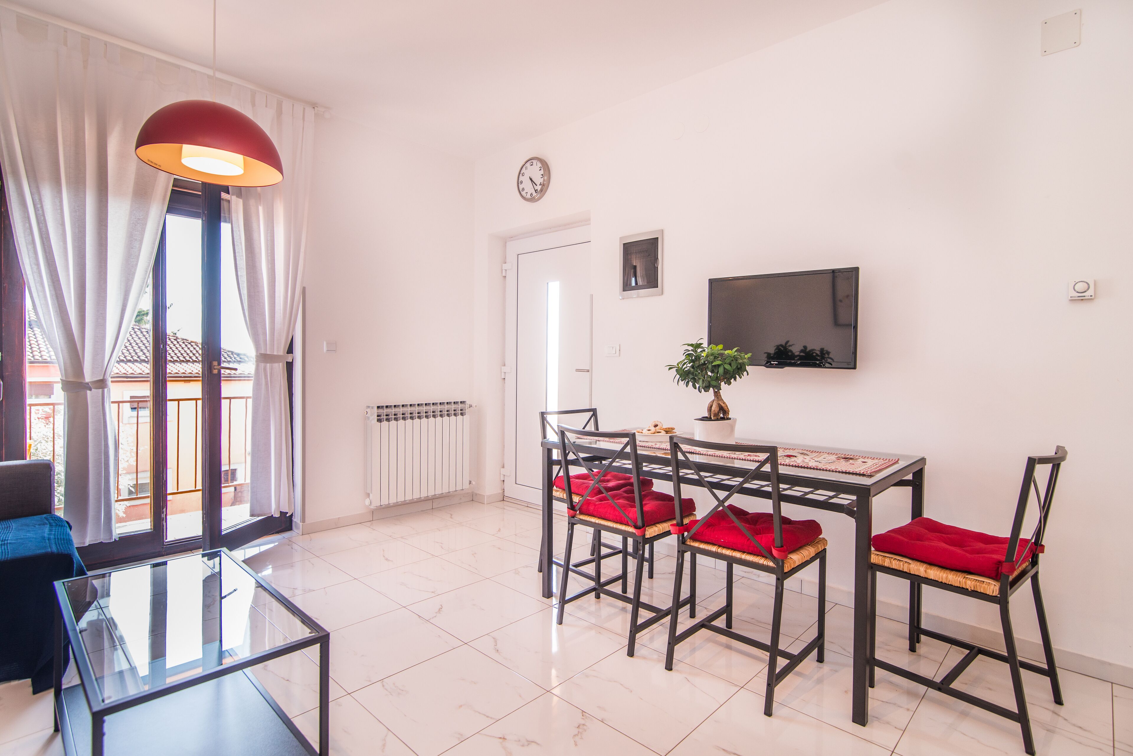 Un coin repas avec une table en verre, quatre chaises dotées de coussins rouges, une plante en pot, un téléviseur fixé au mur et une horloge sur le mur.
