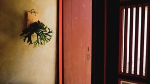Een kamer met een houten deur, een kleine tafel met een waterkoker en een schaal fruit, en een bank met kussens.