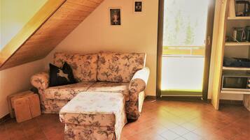 Ein gemütliches Wohnzimmer mit einem blumigen Sofa und Hocker, einer Holzbalkendecke und einer Glastür zum Außenbereich.