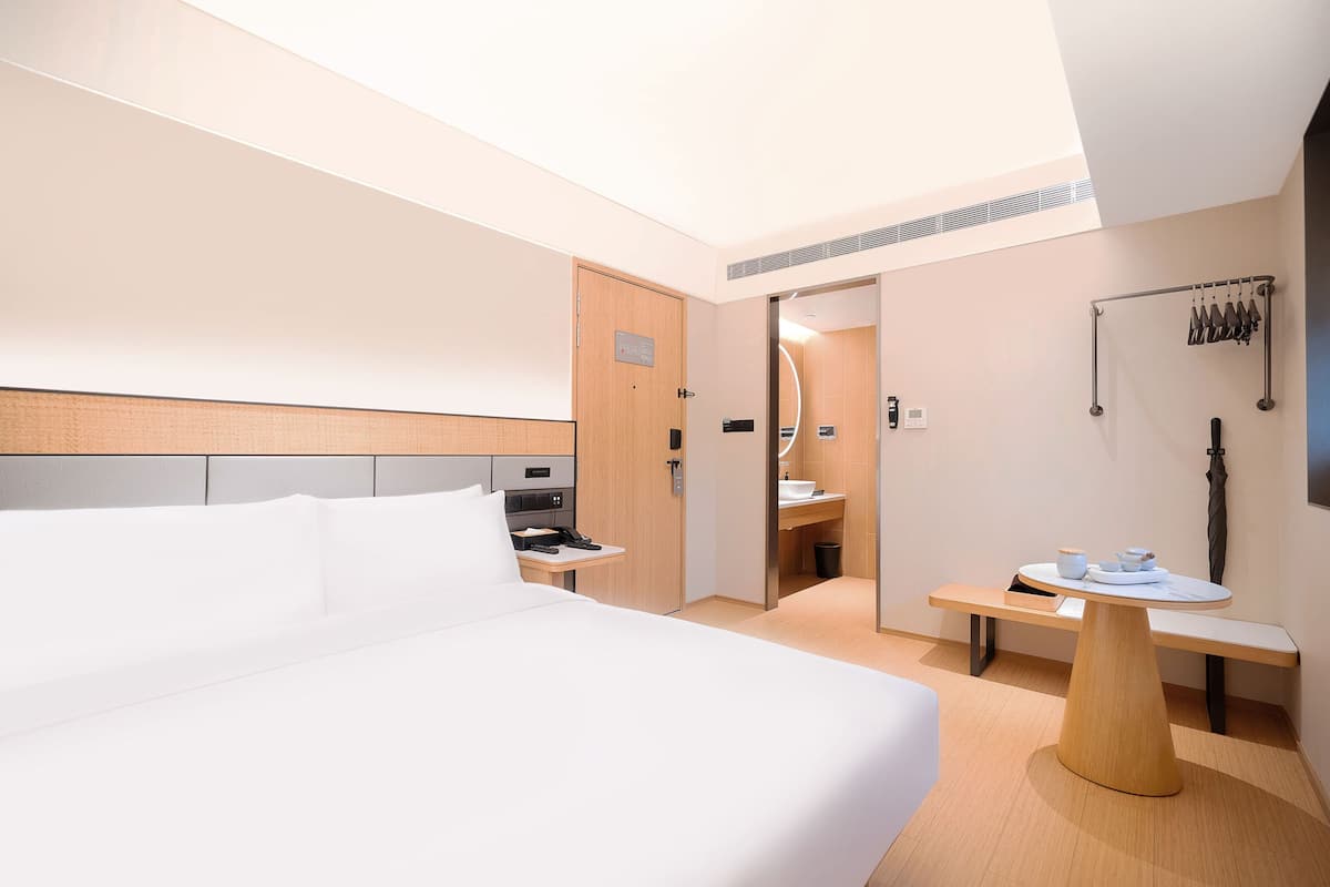 A modern hotel room with a large bed, a bedside table with a phone, a small table with a bowl and cups, a mirror, and a coat rack with hanging items.