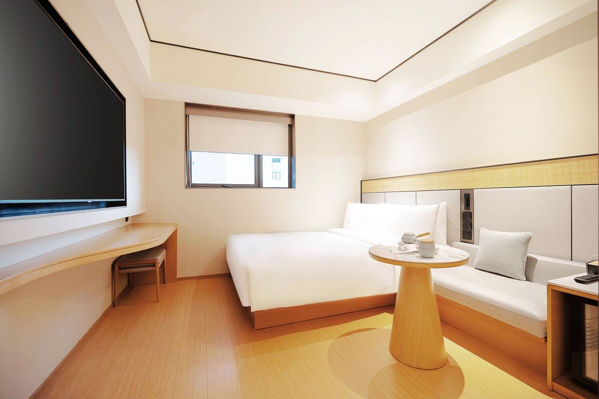A modern hotel room with a bed, a small desk, a round wooden table, and a window.