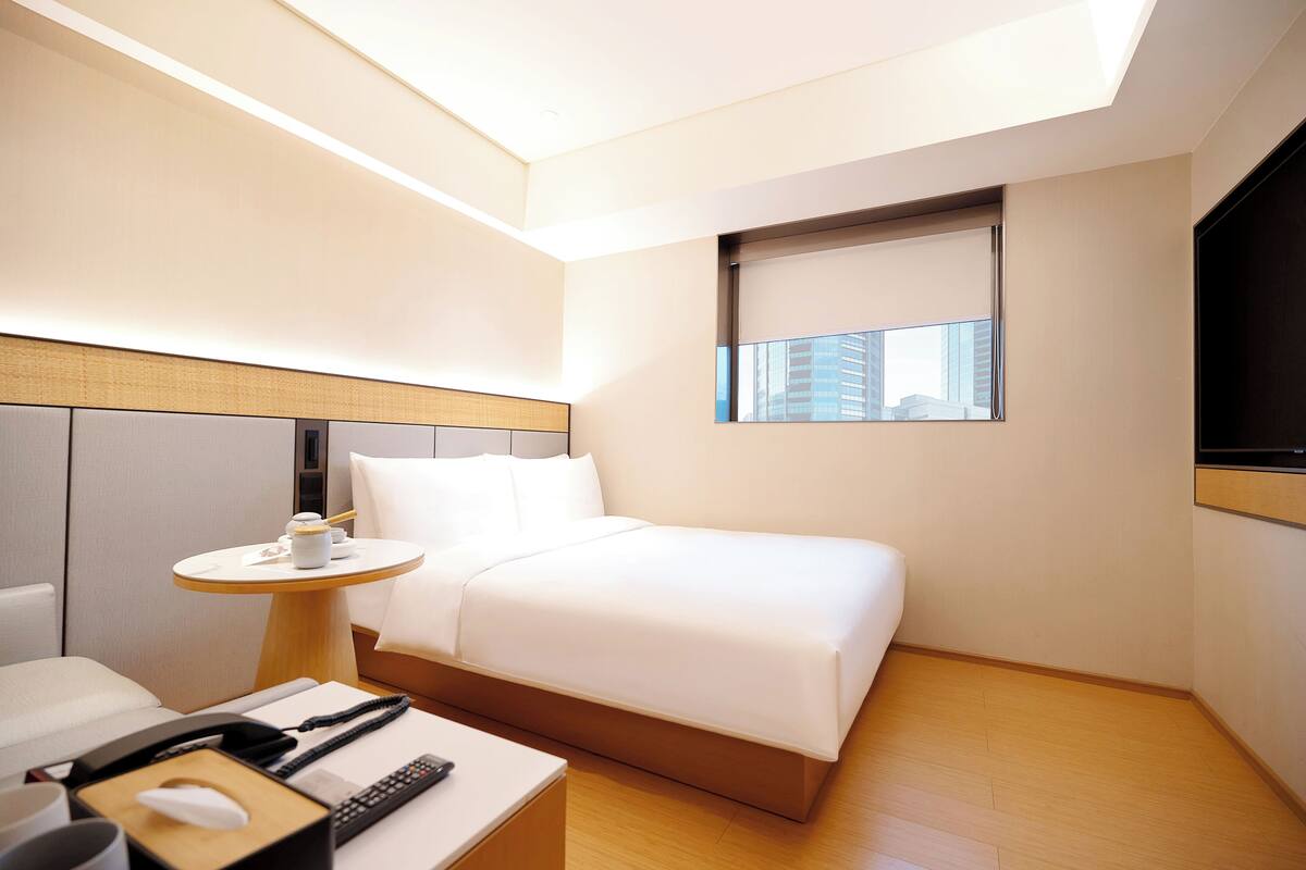 A modern hotel room with a neatly made bed, a bedside table with a lamp, a small table with a cup and a remote control, and a window with a view of buildings.