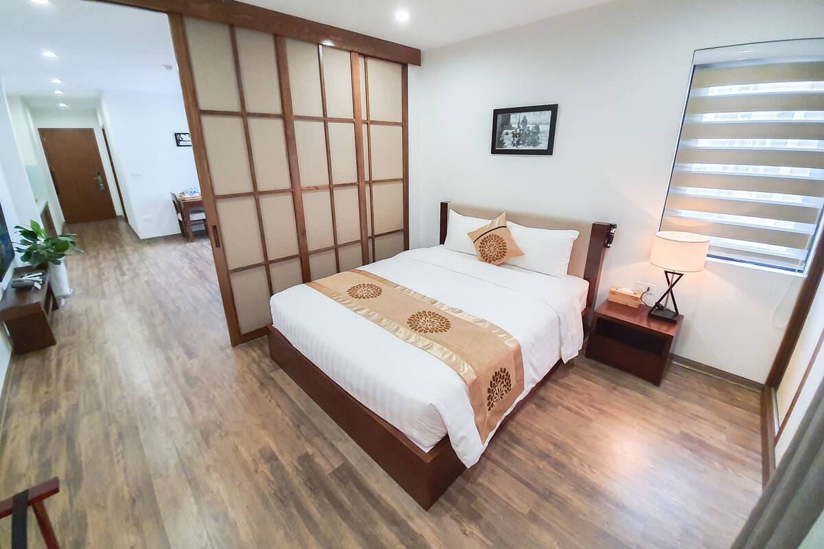 A hotel room with a wooden floor, a bed with white bedding and a decorative throw, a nightstand with a lamp, and a picture on the wall.