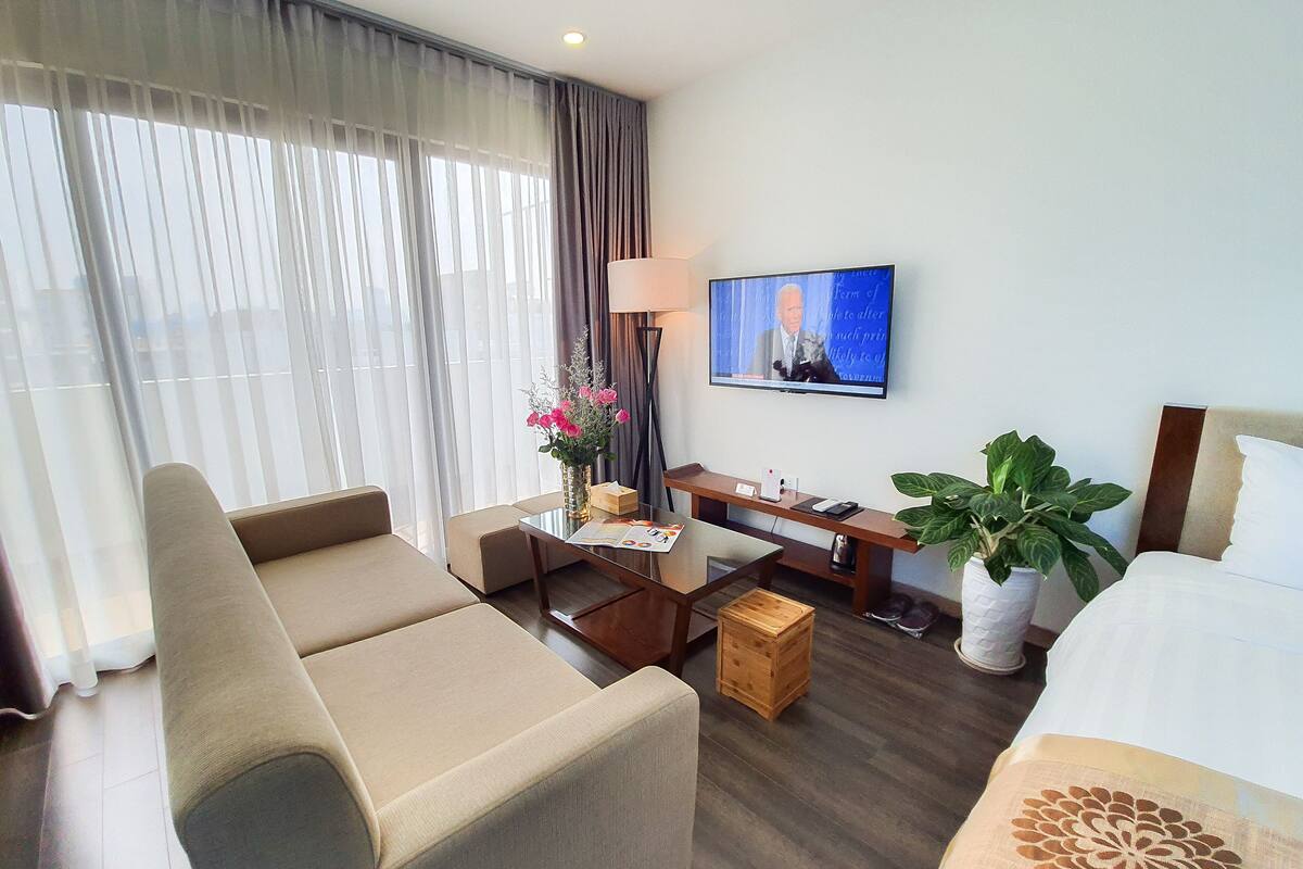 A hotel room with a sofa, a small table with a vase of flowers, a desk with a lamp, a television on the wall, and a bed with a decorative pillow.