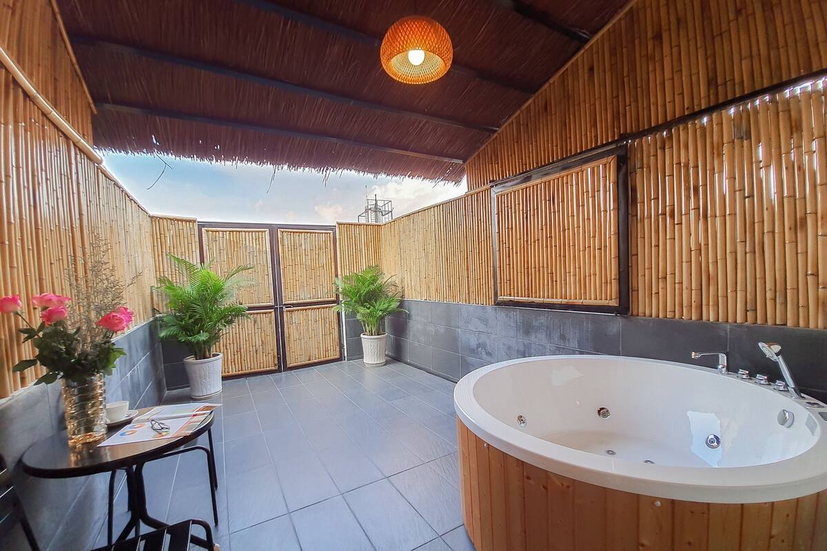 A bathroom with a wooden bathtub, two potted plants, a small table with a chair, and a large window.