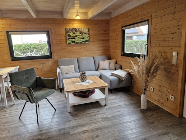 A cozy room with a grey sofa, a wooden coffee table, and a chair. There are two windows with views of greenery outside.