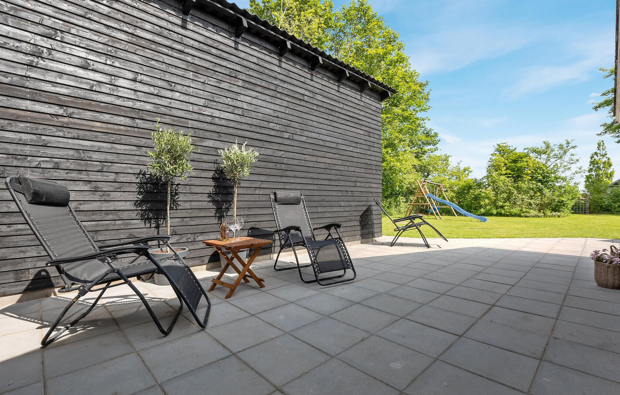 Een terras buiten, met een houten wand, twee klapstoelen, een kleine tafel en een speeltoestel voor kinderen op de achtergrond.