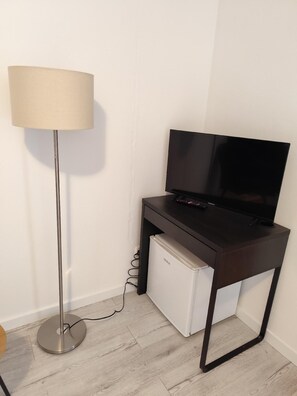 Compact room with a dark desk, flat-screen TV, mini-fridge, and floor lamp.
