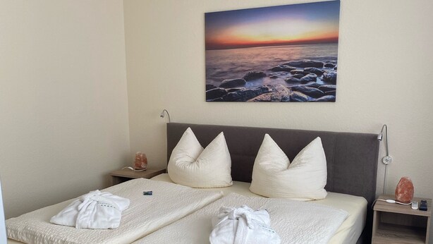 A neatly made bed with white linens and towels, a grey headboard, and a painting of a sunset over the ocean on the wall.