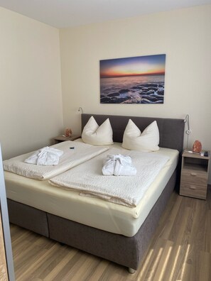 A neatly made bed with white linens and towels, a grey headboard, and a painting of a sunset over the ocean on the wall. - Apartment "Sandra" (Heilbad Heiligenstadt)