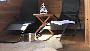 Interior de una cabaña de madera con dos sillones reclinables, una mesita con un modelo de barco de vela, una alfombra de piel y un cubo de metal.