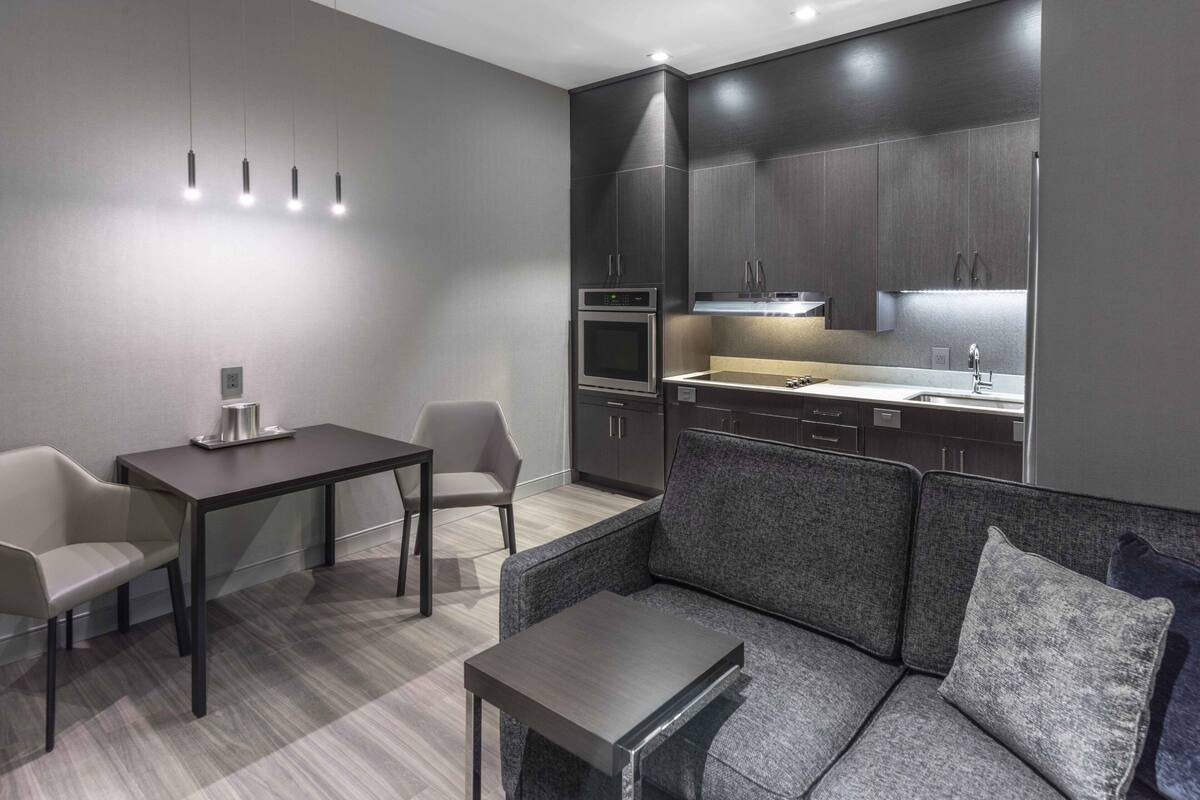 A modern kitchen with dark cabinetry, a built-in oven, and a countertop with a sink. There is a dining area with a table and chairs, and a seating area with a sofa and a coffee table.