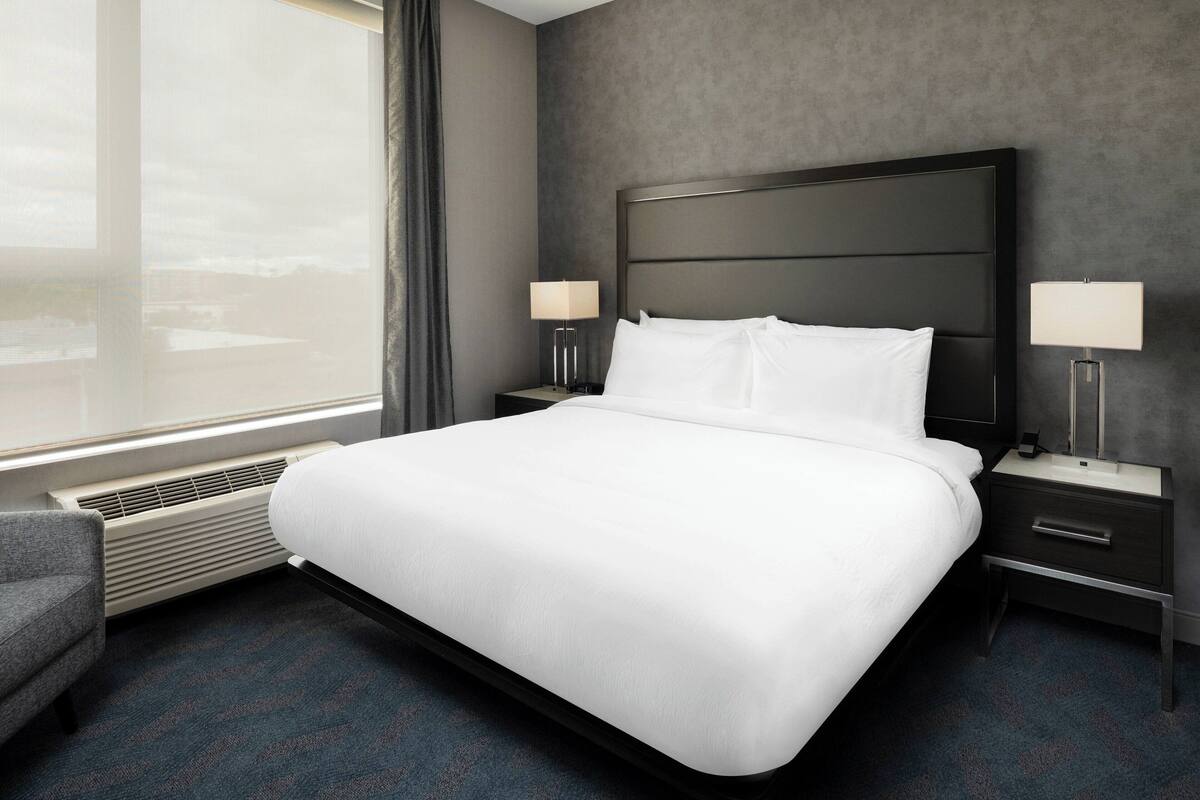 A hotel room with a large bed, two bedside tables with lamps, a chair, and a view of the city through the window.