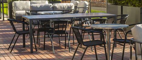 Un patio con mobiliario exterior, incluyendo mesas y sillas, rodeado de suelo de ladrillo y follaje.