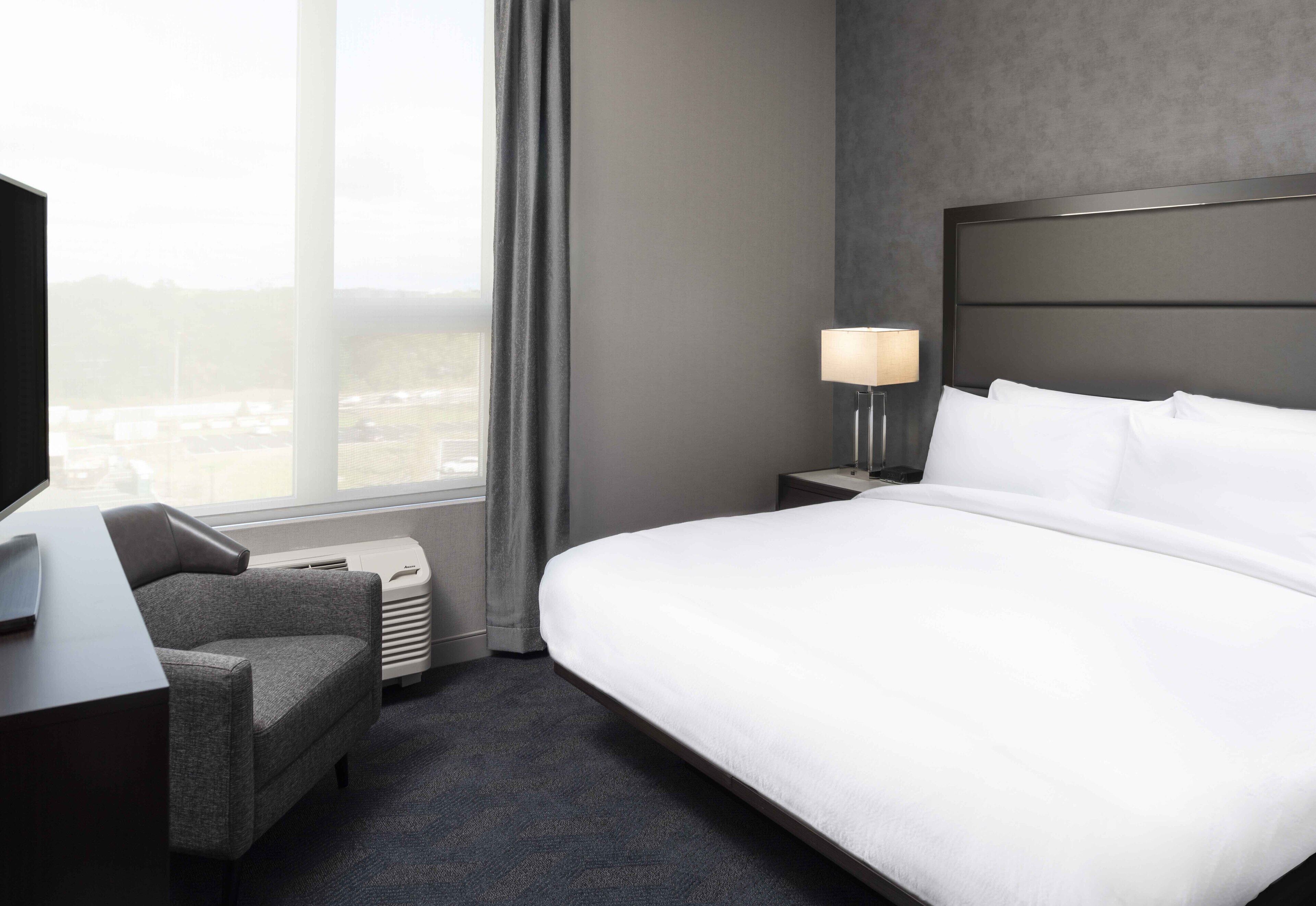 A hotel room with a large bed, a desk with a lamp, a chair, and a view of the city through the window.