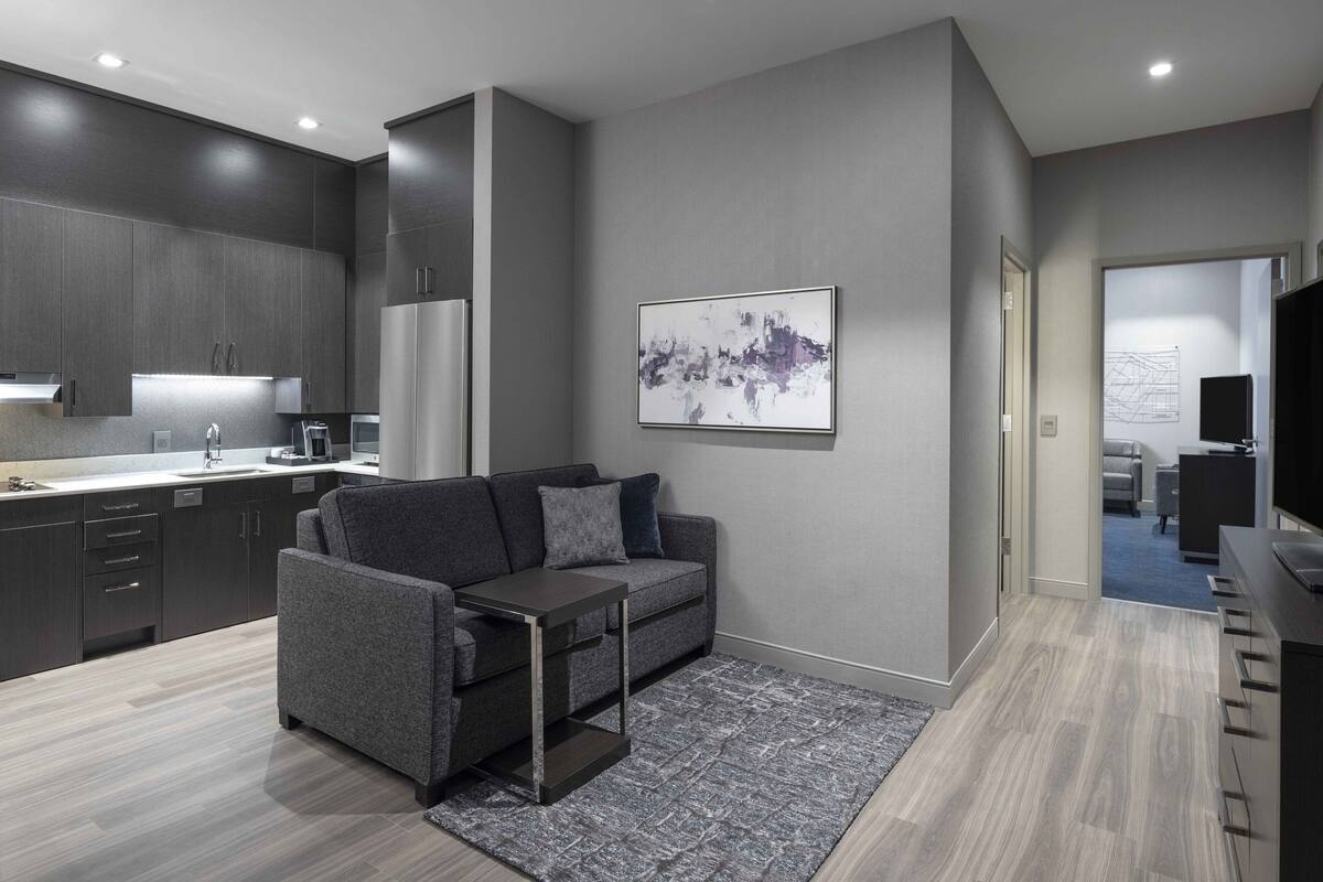 A modern living room with a grey sofa, a coffee table, and a kitchen area with dark cabinets and a refrigerator.