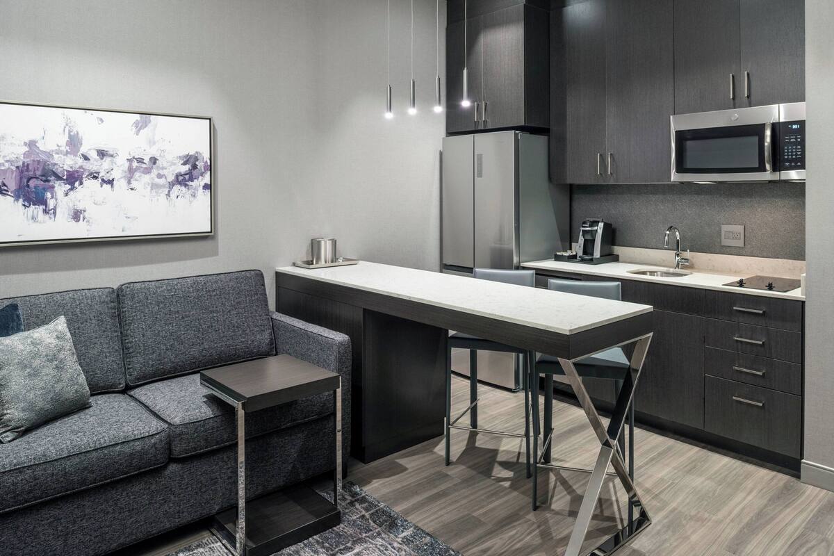 A modern kitchen with dark cabinetry, a stainless steel refrigerator, a microwave, and a bar area with stools.