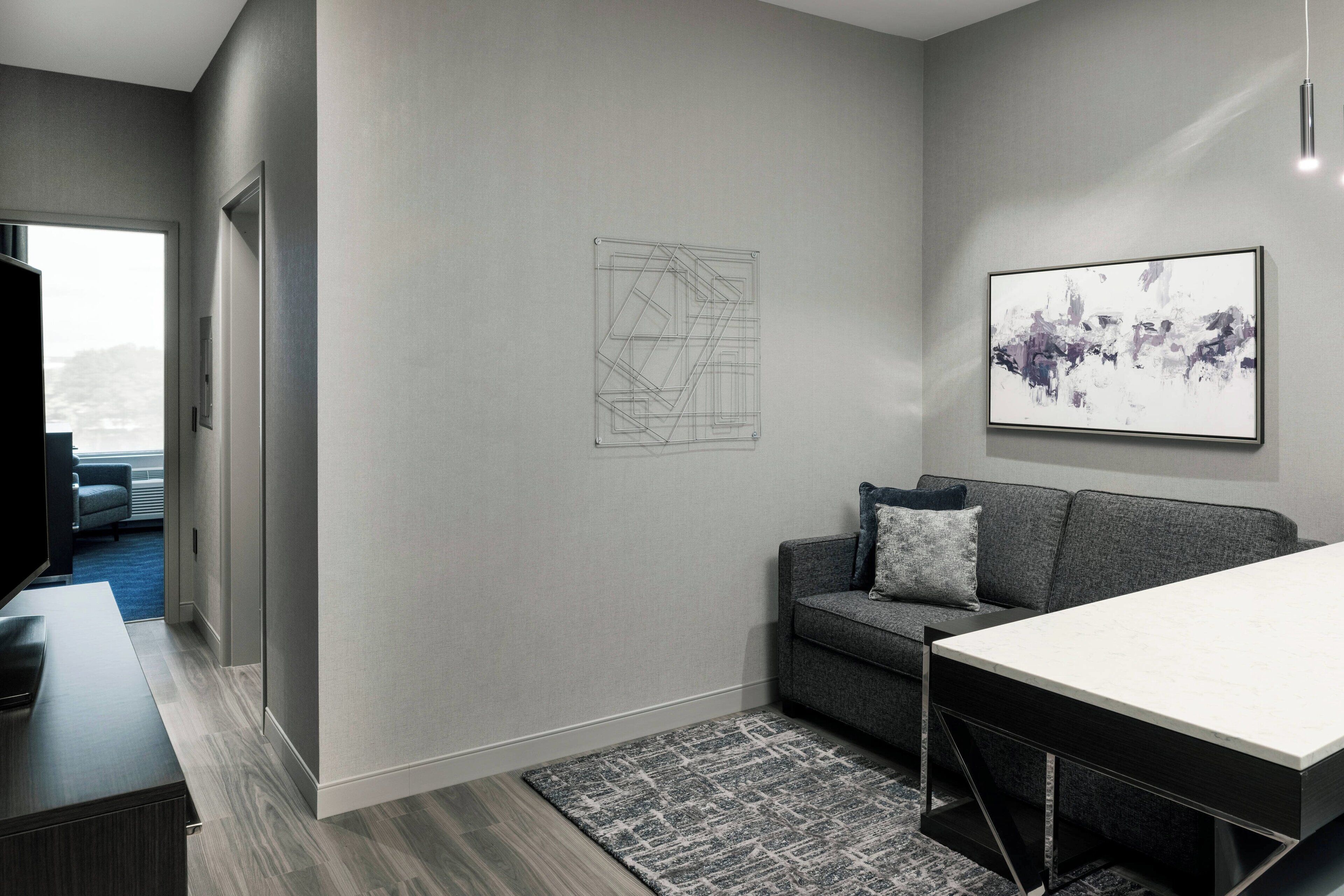 A modern living room with a grey sofa, a coffee table, and a rug. There is a framed abstract artwork on the wall and a door leading to another room.