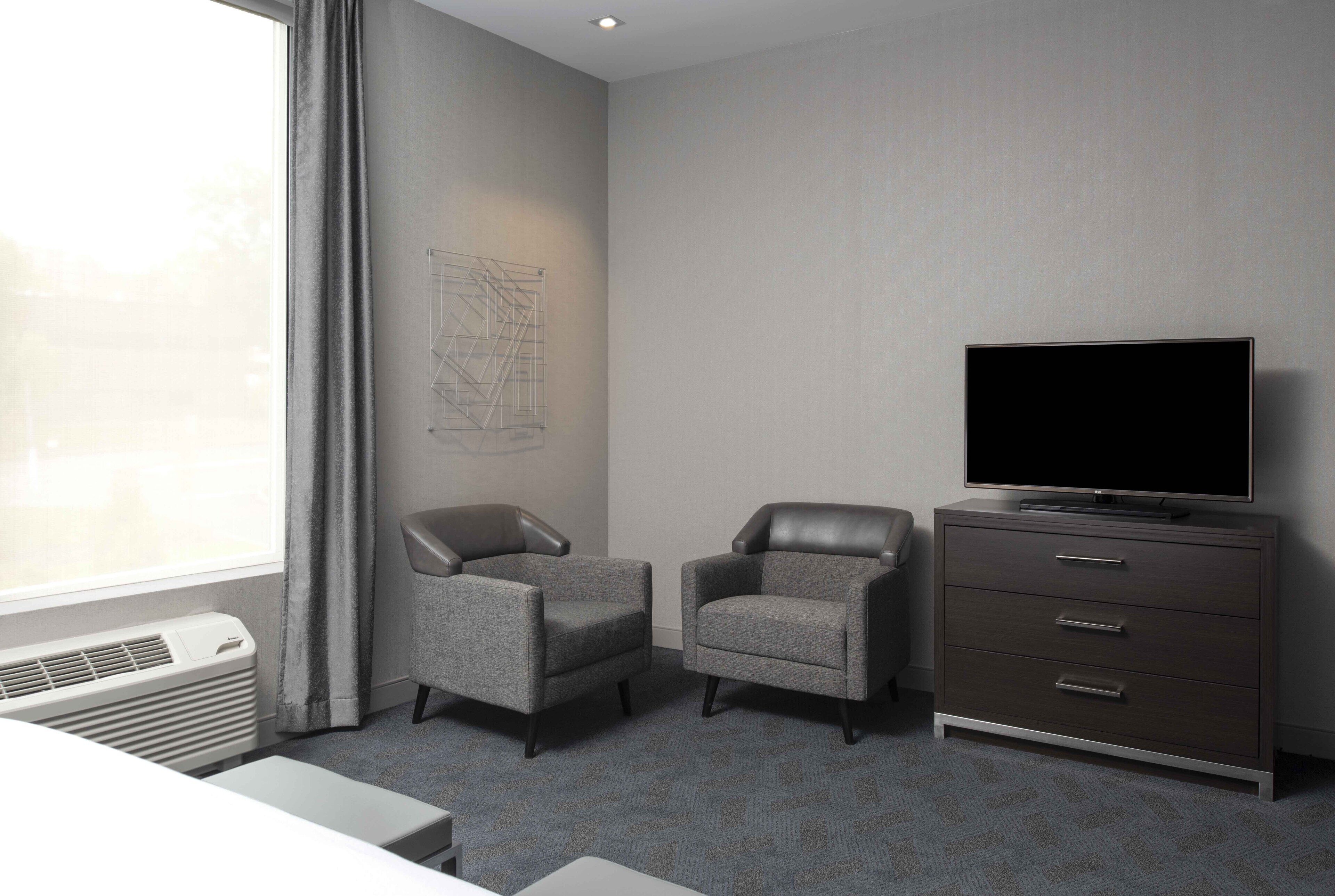 A hotel room with two grey armchairs, a dark wooden dresser with a television, and a window with curtains.