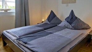 A double bed with a striped bedspread and two pillows, a nightstand with a lamp, a window with a view of a beach, and a painting of a beach scene on the wall. - Wide view - ideally located family apartment with charm (Wangerooge)