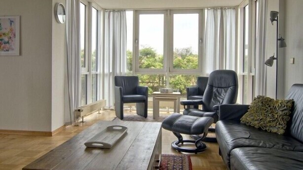 A spacious living room with a large window, a black leather sofa, two armchairs, a coffee table, and a small table with a lamp. - Wide view - ideally located family apartment with charm (Wangerooge)
