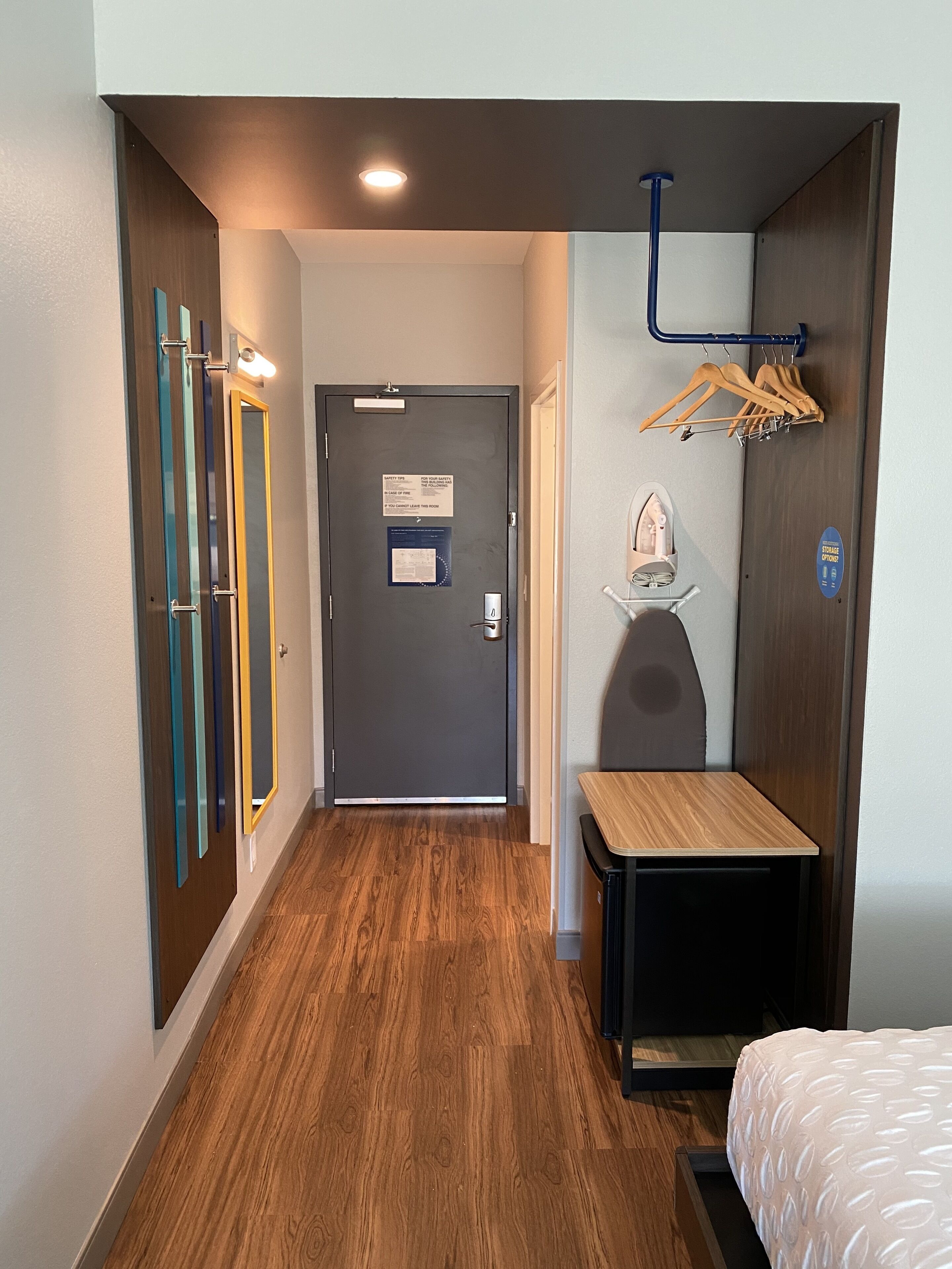 Um corredor de hotel com piso de madeira, uma porta com sinal de segurança, um porta-roupas com cabides e uma pequena mesa com uma cadeira.