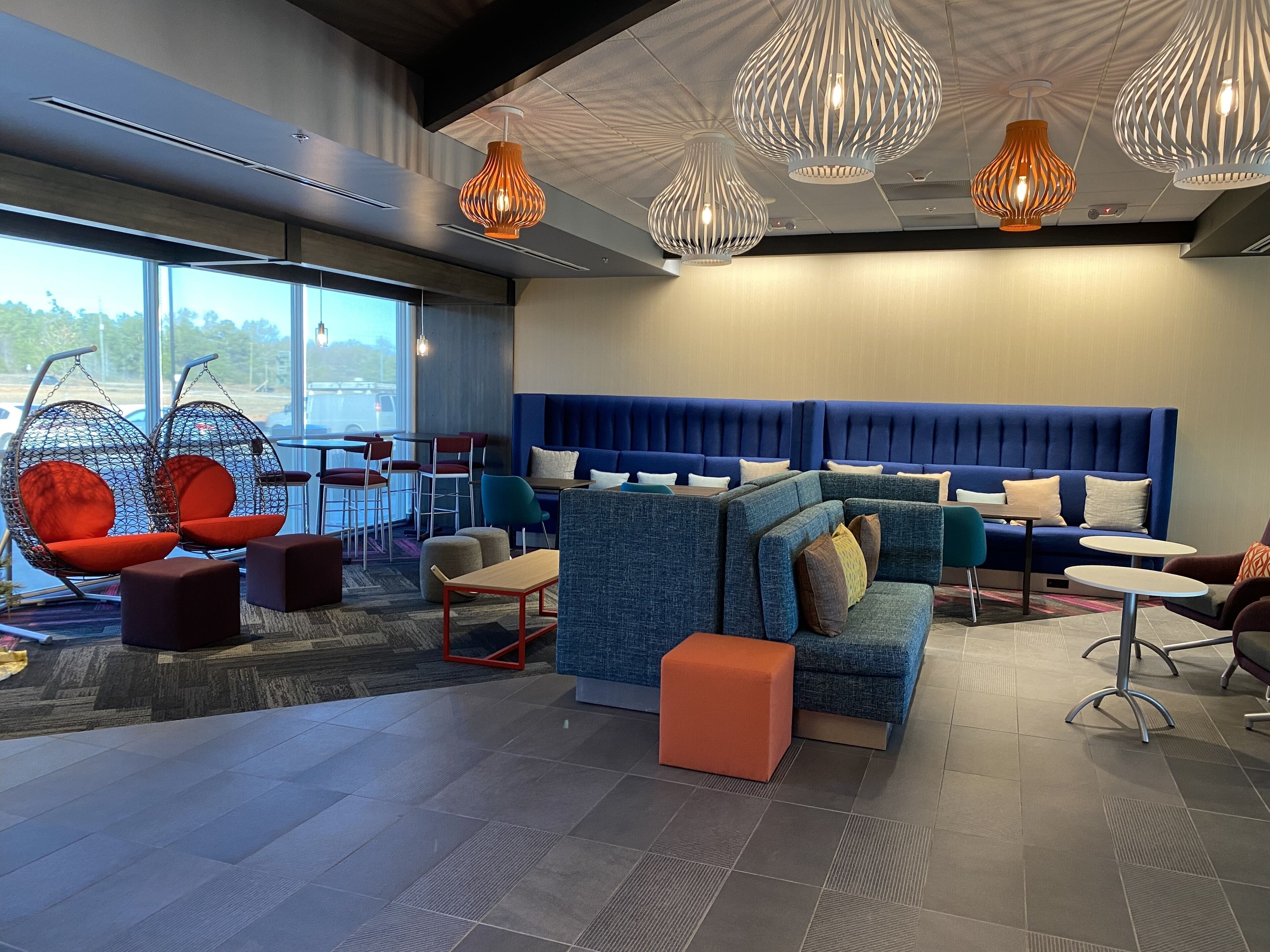 A modern lounge area with seating arrangements, including a blue sofa and red chairs, and a large window providing a view outside.