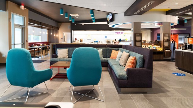 Un vestíbulo moderno con un sofá modular, sillas color turquesa, una barra de café y un área de restaurante visible.