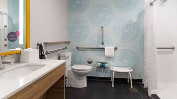 Un baño moderno con lavamanos, inodoro y zona de ducha blancos. Hay un pasamanos y un taburete pequeño cerca del inodoro.