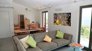 A modern living room with a grey sofa, green and beige cushions, a glass coffee table, and a dining area with a wooden table and chairs.