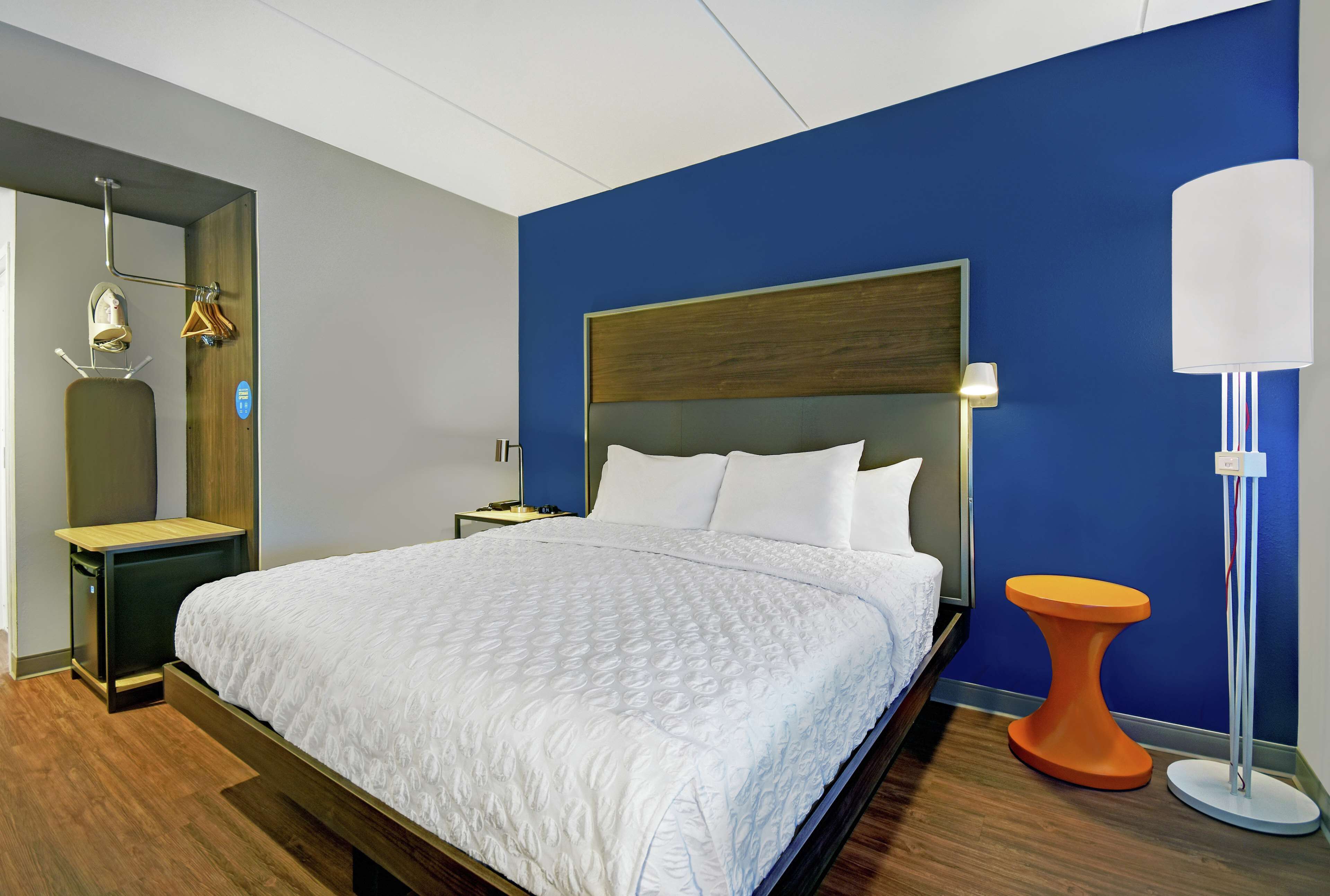 Una habitación de hotel moderna con una cama grande, una mesita de noche, una lámpara y un taburete pequeño naranja.