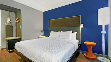 Una habitación de hotel moderna con una cama grande, una mesita de noche, una lámpara y un taburete pequeño naranja.