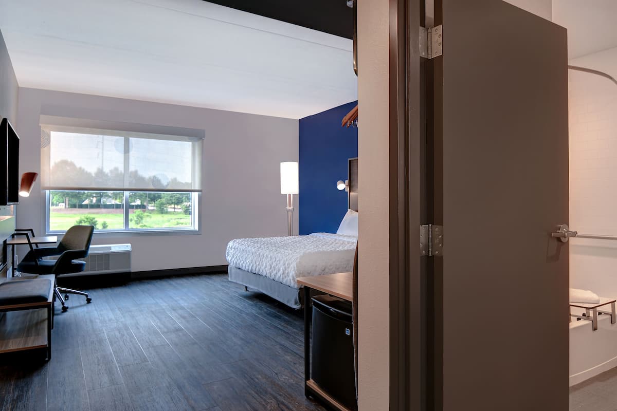 A hotel room with a bed, desk, chair, and a view of a green landscape through a large window.
