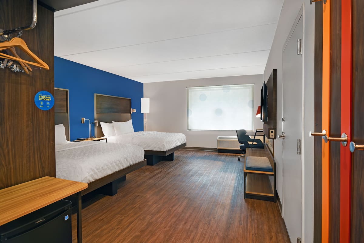 A hotel room with two beds, a desk, a chair, a TV, and a window with blinds.