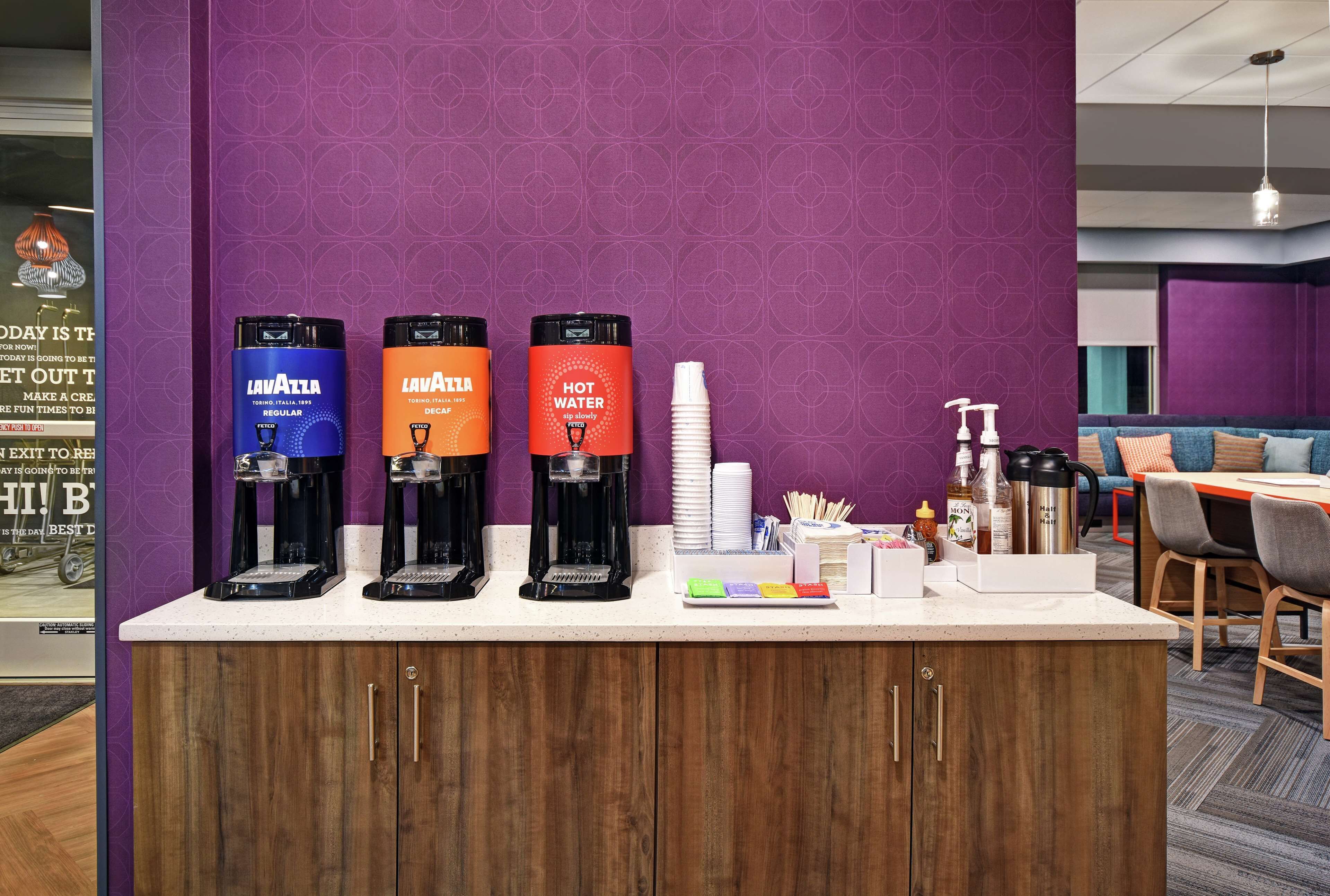 A coffee station with three dispensers labeled 'Lavazza' and 'Hot Water' in a modern room with purple walls and wooden cabinets.