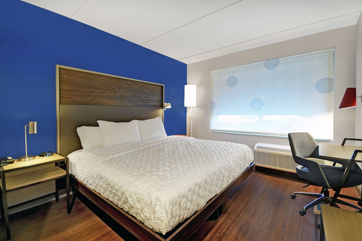 A hotel room with a large bed, a desk with a chair, a nightstand, and a window with blinds.