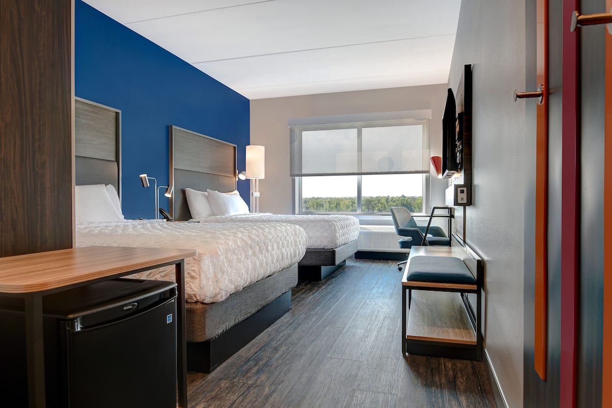 A hotel room with two beds, a desk, a chair, a TV mounted on the wall, and a window offering a view of the outdoors.
