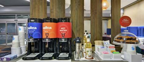 En kaffestation med tre kaffebryggare märkta med "Lavazza" och "Varmt vatten", en kaffekanna samt ett urval av tillbehör och drycker.